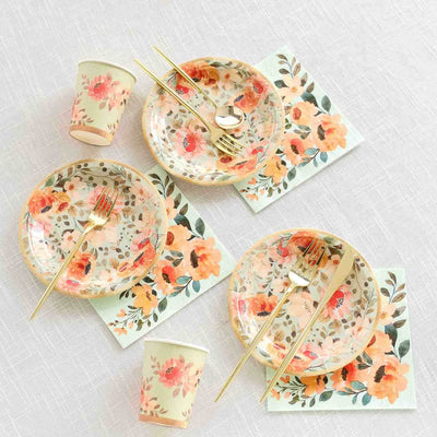 Floral dinnerware set with gold utensils and paper cups on a textured tablecloth.