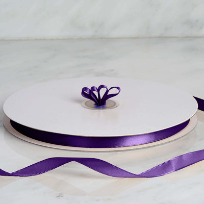 White ribbon spool with purple satin ribbon and bow center on marble surface