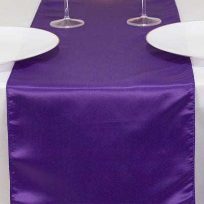 Purple satin table runner on a white table with plates and glasses.