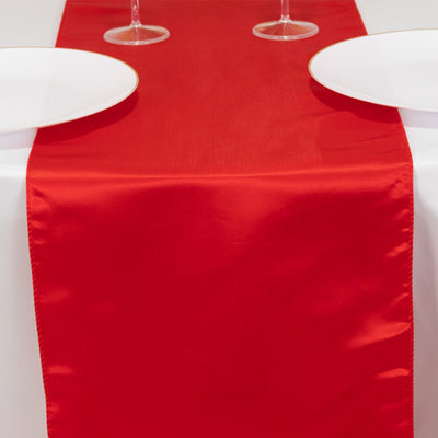 Red satin table runner on a white table with plates and glasses.