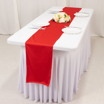 Red table runner on a white table setting with plates and flowers.