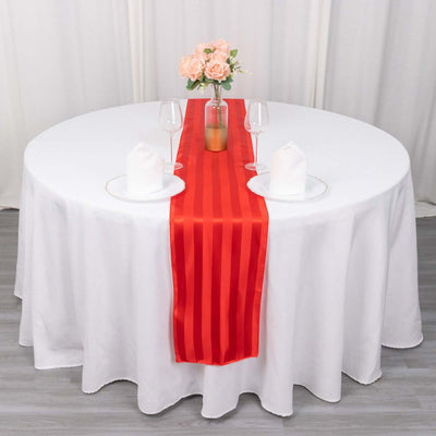 Red satin table runner on a round white table set for dinner with flowers and glasses.