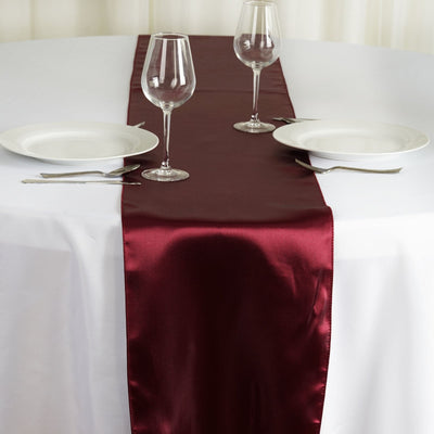 Burgundy satin table runner on a white table with glasses and plates.