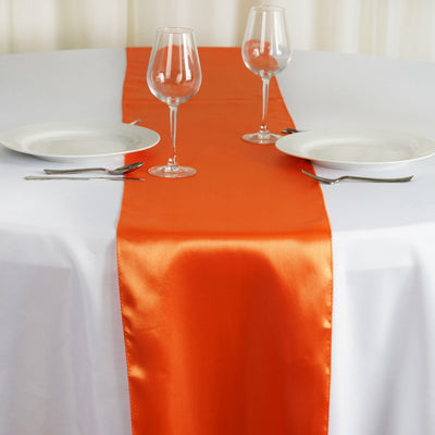 Orange satin table runner on a white table setting with plates and glasses.