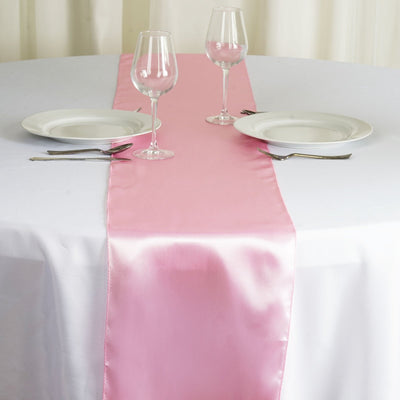 Pink satin table runner on a white table with dinner plates and glasses.