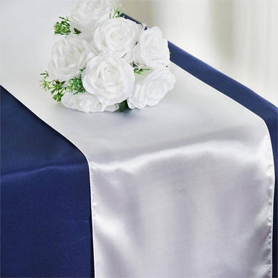 White satin table runner with artificial roses on a navy tablecloth.