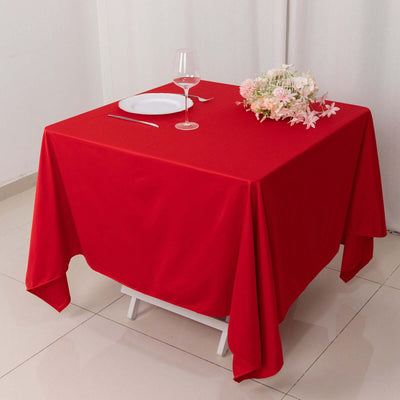 Red tablecloth draped over a square table set with a plate, glass, and flowers.