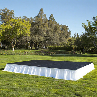 Outdoor event stage with black surface and white skirt in a grassy setting.
