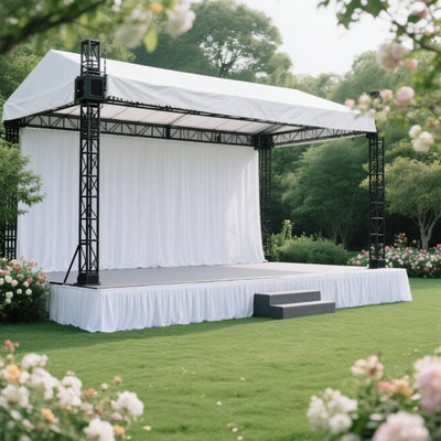 Outdoor event stage with white drapery and black framework, surrounded by greenery.
