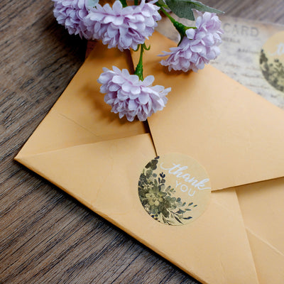 Yellow envelope with floral thank you sticker and purple flowers on wooden surface.