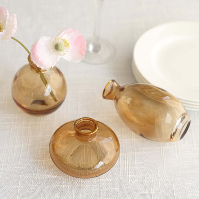 Set of three amber glass vases with flowers and plates in the background.