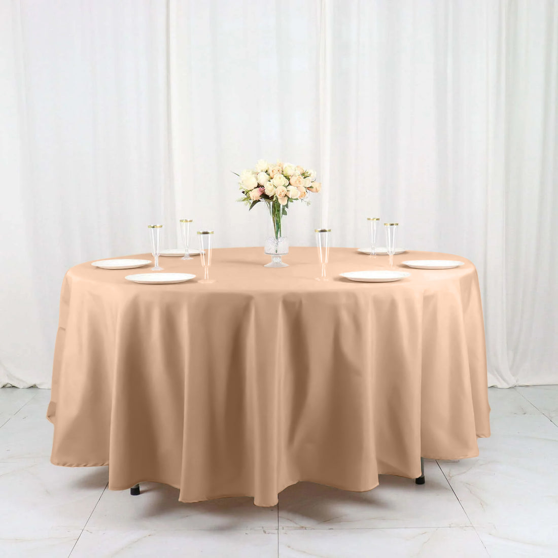 Round beige tablecloth draped over a table with plates and floral centerpiece.