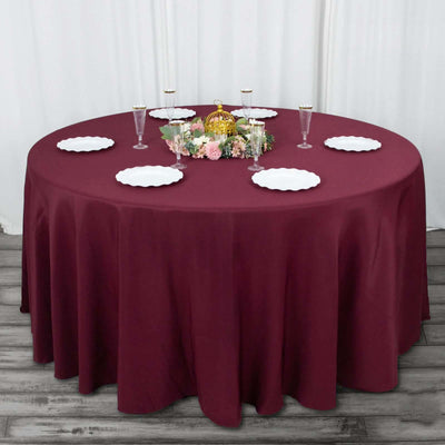 Round burgundy tablecloth on a round table set for dining with plates and floral centerpiece.