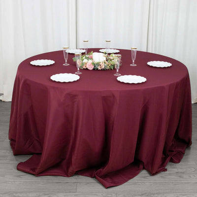 Round burgundy tablecloth on a dining table set with plates and floral centerpiece.