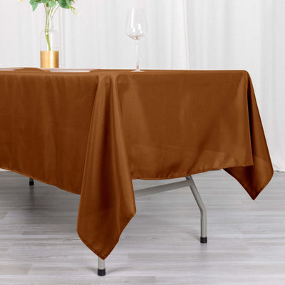 Brown tablecloth draped over a table with a glass and plant in the background.