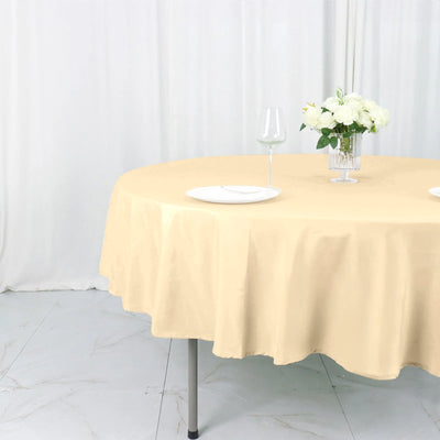 Round beige tablecloth draped over a table with plates and a vase of flowers.