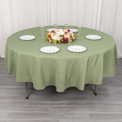 Round olive green tablecloth on a dining table set for a meal with plates and flowers.
