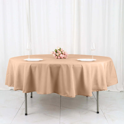 Round beige tablecloth on a table with plates and flowers.