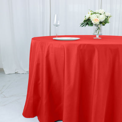 Red tablecloth draped over a round table with a glass and vase of flowers.