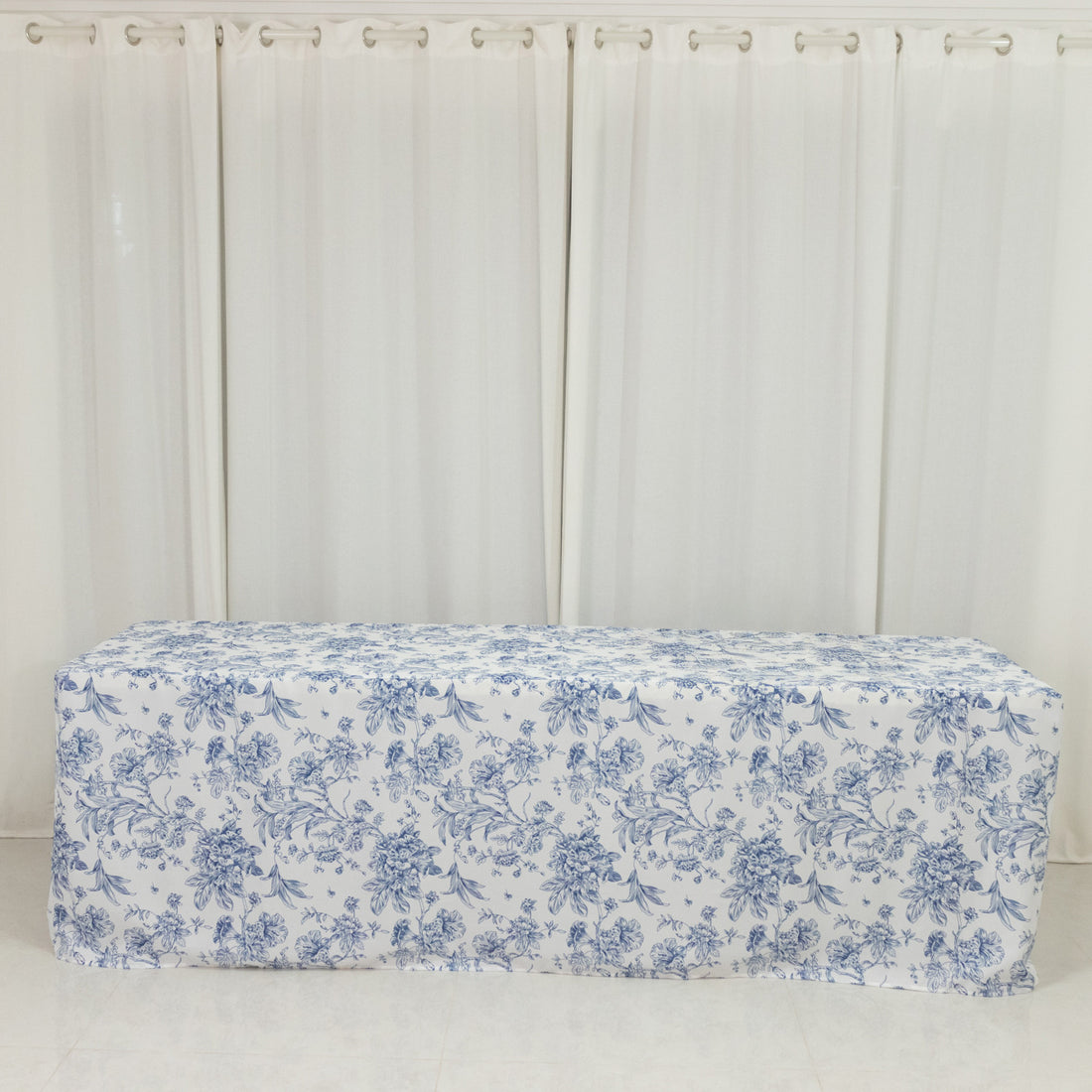 Floral patterned tablecloth draped over a table against white curtains.