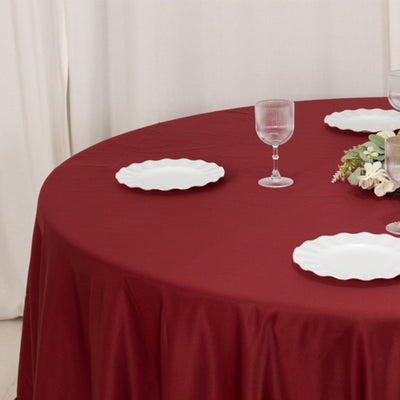 Burgundy round tablecloth with white plates and glassware on a dining table.