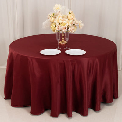 Round maroon tablecloth draped over a table set with plates and a floral centerpiece.
