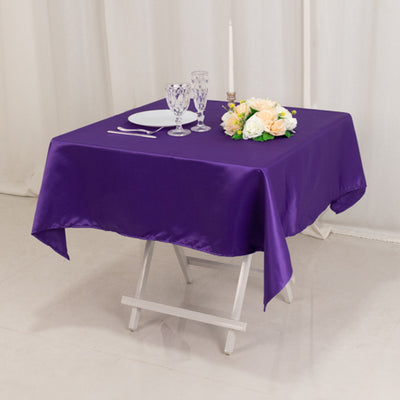 Purple satin tablecloth on a folding table with decorative flowers and glassware.