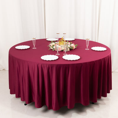Round burgundy tablecloth draped over a table with plates and floral centerpiece.