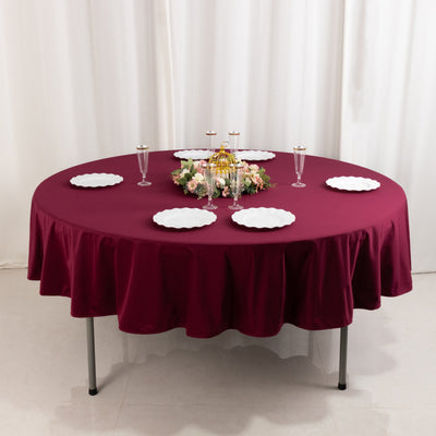 Round burgundy tablecloth set with white plates and floral centerpiece.