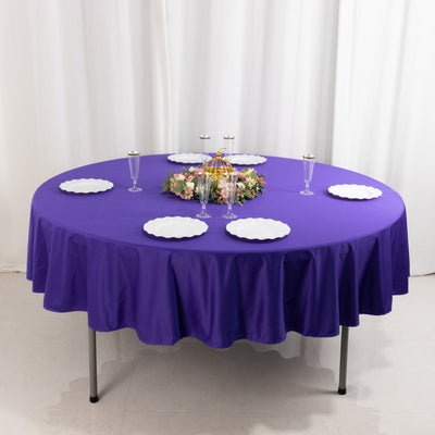 Purple round tablecloth with white plates and floral centerpiece on a table.