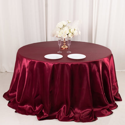 Burgundy satin tablecloth draped over a round table with plates and floral centerpiece.