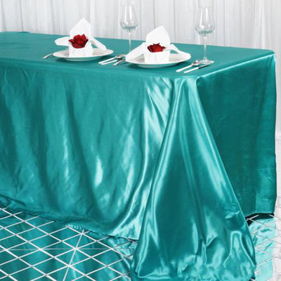 Teal satin tablecloth with plates and rose centerpieces on a patterned floor.
