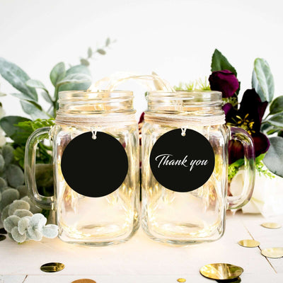 Two clear mason jars with "Thank you" tags, surrounded by greenery and decor.