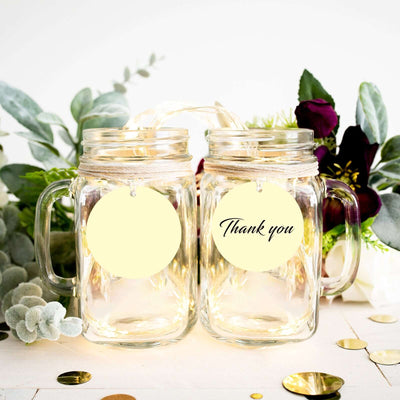 Two glass mason jars with "Thank you" tags surrounded by greenery and decorative elements.
