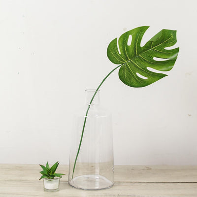 Green monstera leaf in a clear glass vase on a wooden surface.
