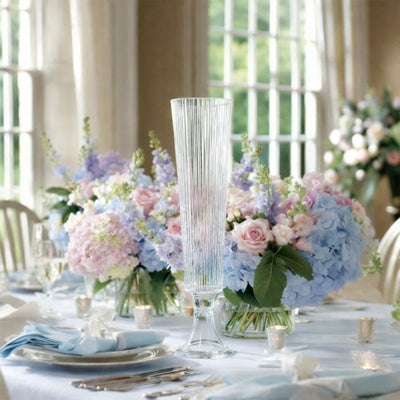 Elegant tall glass vase surrounded by pastel floral arrangements on a dining table.