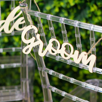 Wooden "Groom" sign on clear chair against greenery backdrop.