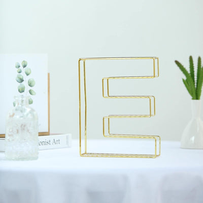 Gold wire letter "E" decorative piece on a table with greenery in the background.