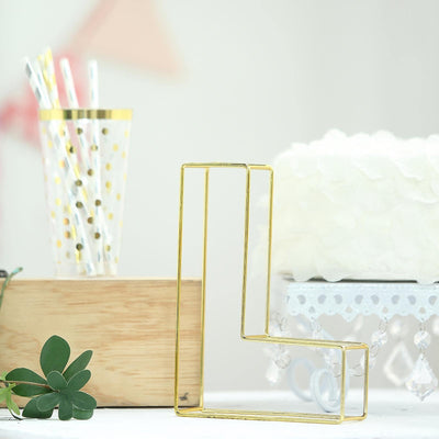 Gold geometric letter "L" decor piece on a table with plants and cake.