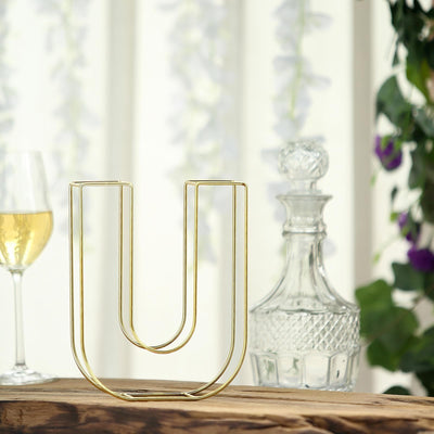 Gold wire vase in U-shape design on wooden table with glassware in the background.
