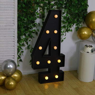 Large black number 4 marquee light with bulbs, surrounded by greenery and balloons.