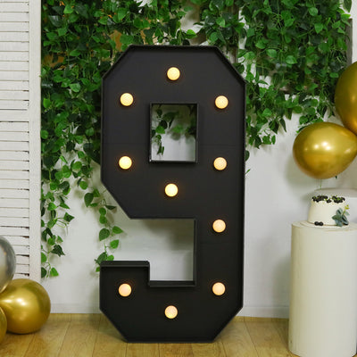 Large black marquee number 9 with lights, surrounded by greenery and gold balloons.