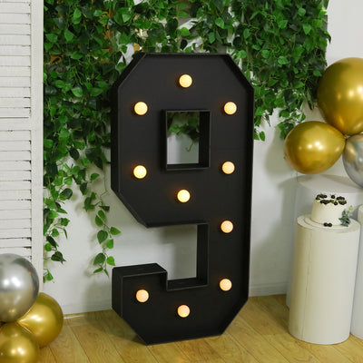 Large black marquee number 9 with lights, set against greenery and balloons.