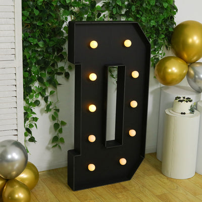 Decorative black marquee letter "D" with lights, surrounded by greenery and balloons.