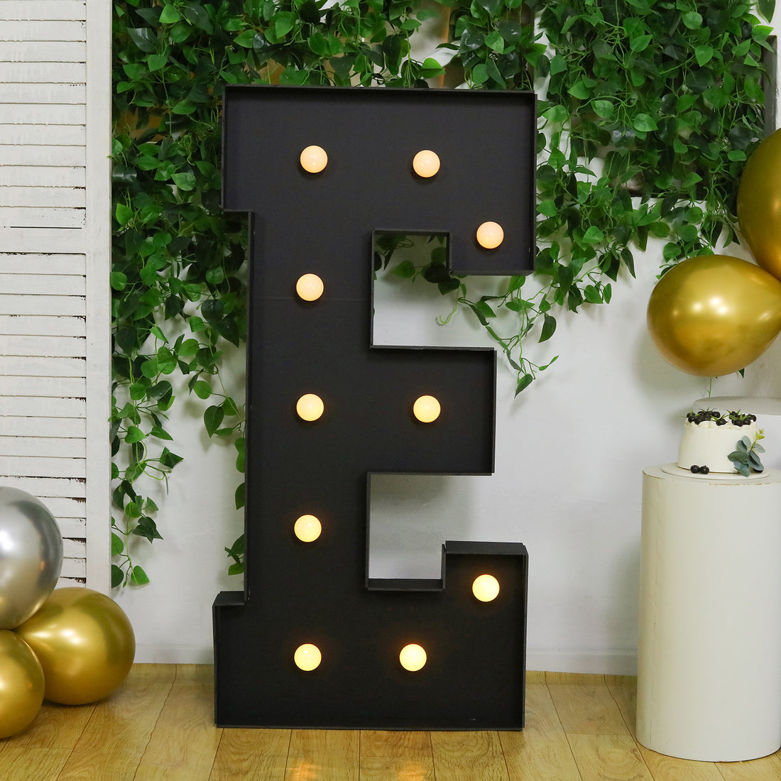 Large illuminated letter 'E' on a backdrop of greenery and balloons.
