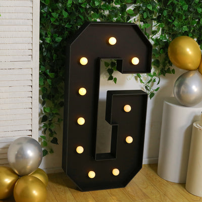 Large illuminated letter G with bulbs, surrounded by greenery and balloons.
