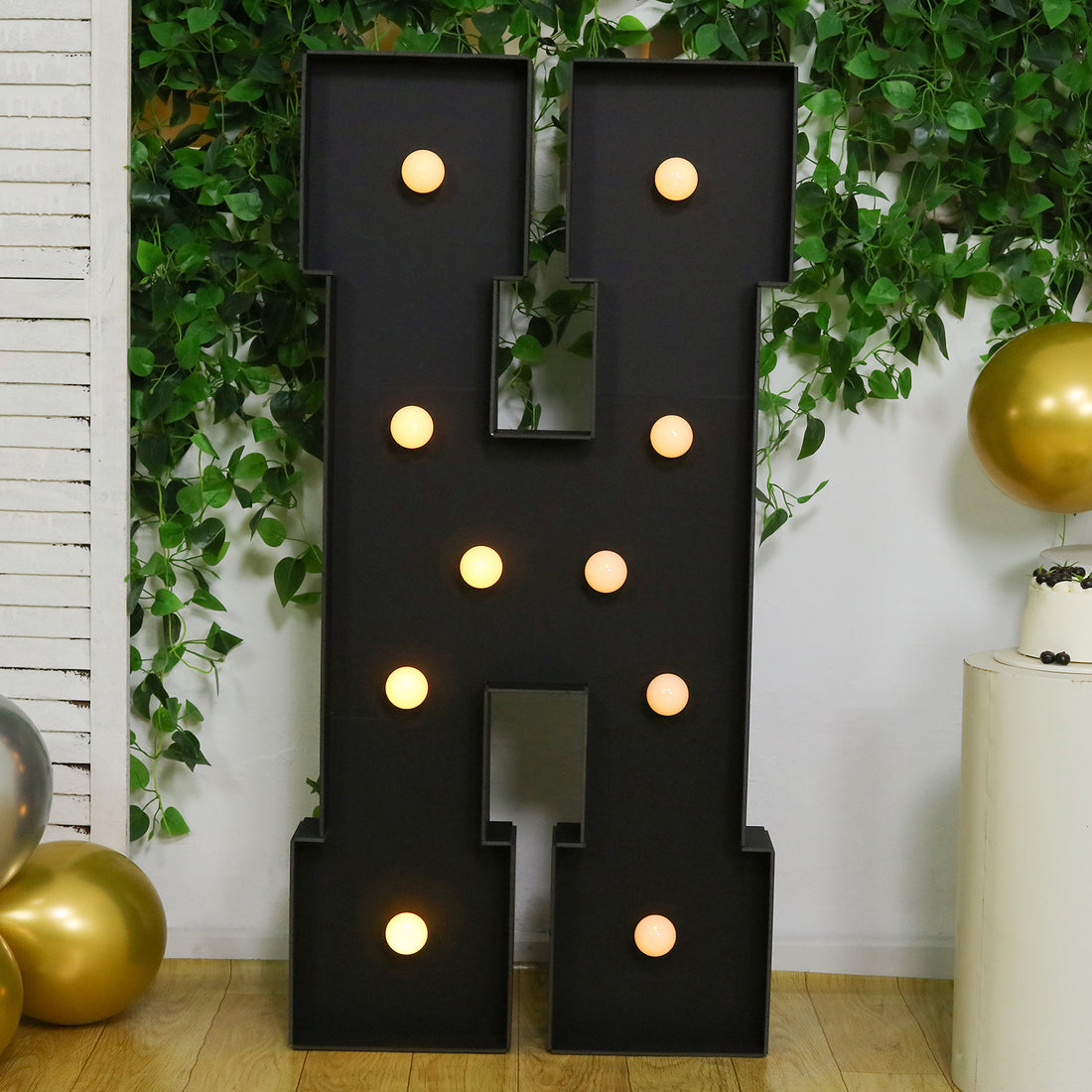 Black marquee letter "H" with lights, set against greenery and balloons.