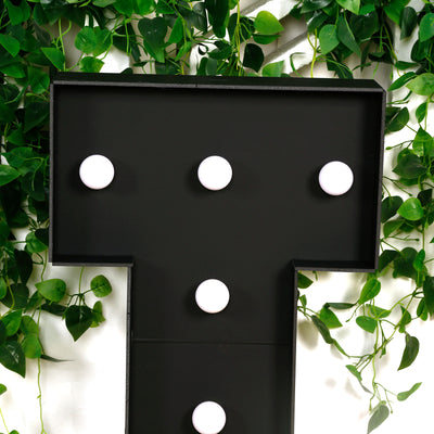 Black marquee letter "T" with bulbs against green foliage.