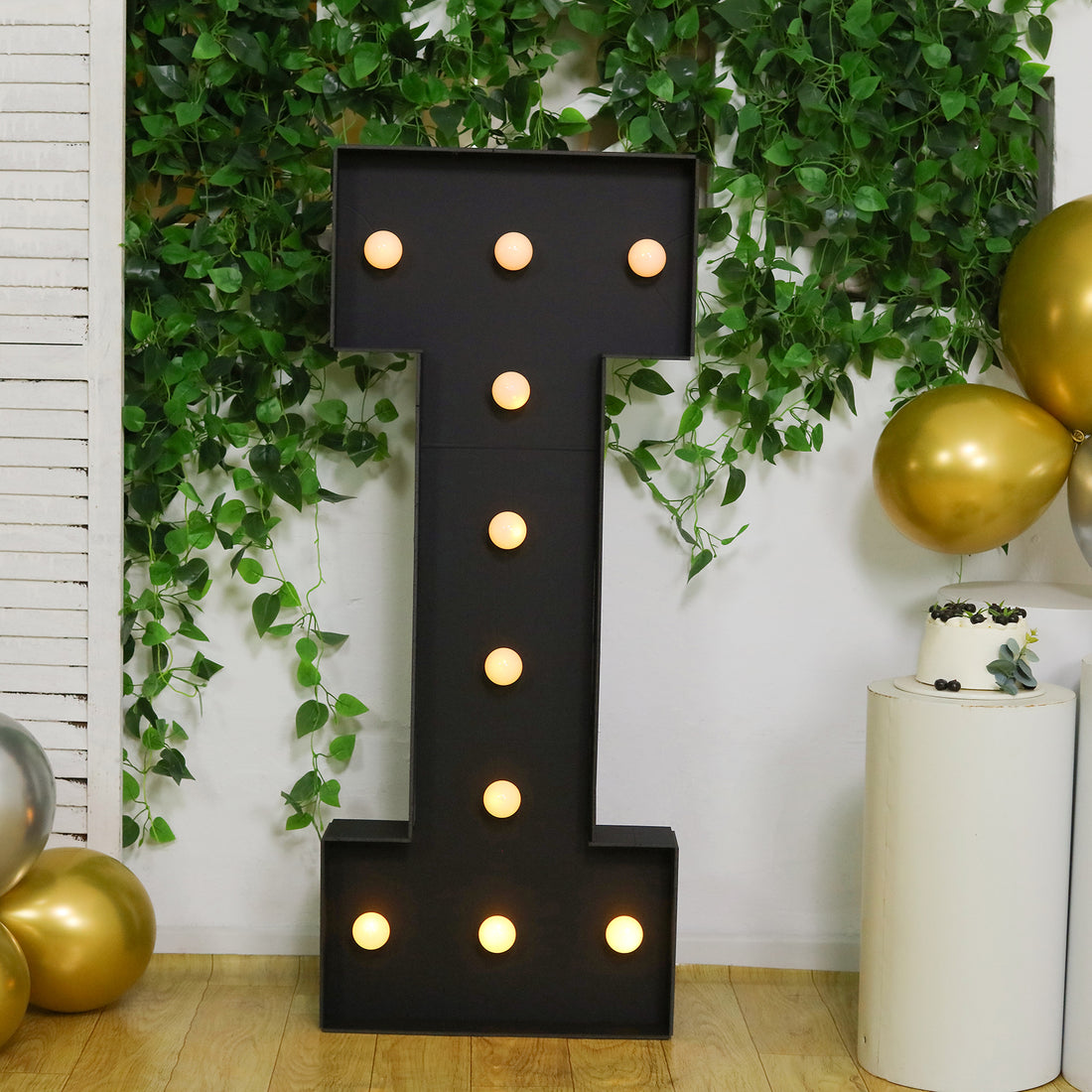 Black marquee letter "I" with lights, surrounded by greenery and gold balloons.