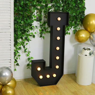 Large black letter 'J' marquee sign with lights, surrounded by greenery and balloons.